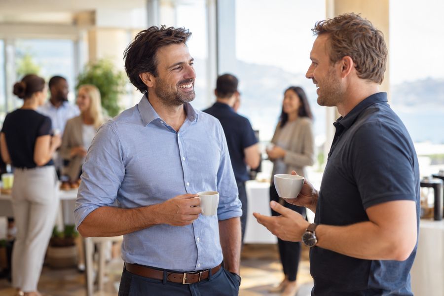 Dois empresários conversam e sorriem, com café na mão, em um evento de negócios